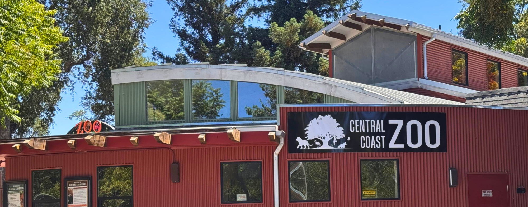 Front view of the Central Coast Zoo’s main building with the zoo’s logo on the sign, framed by tall trees and a bright blue sky in the background.
