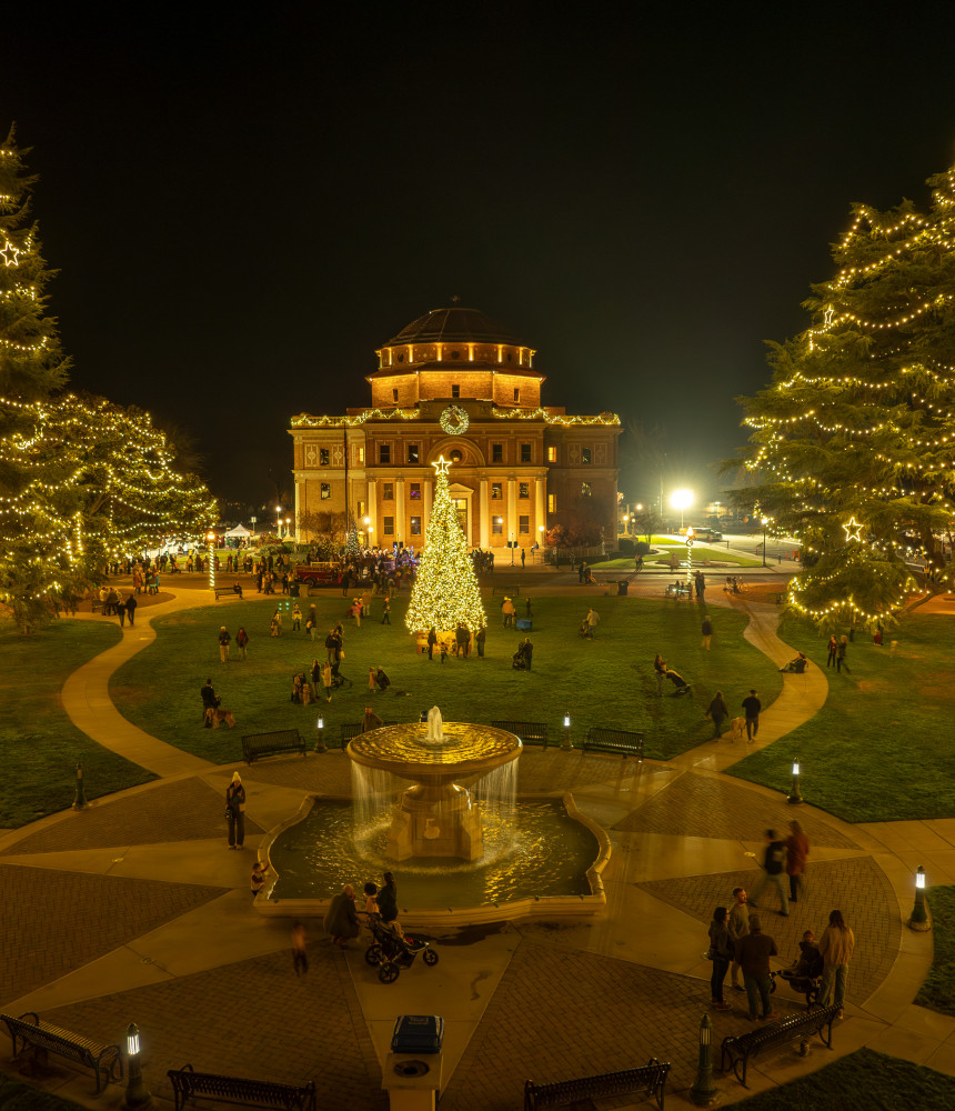 Atascadero’s Sunken Gardens and Historic City Hall festively illuminated with warm glowing Christmas lights, as people stroll through the park and around the building. 