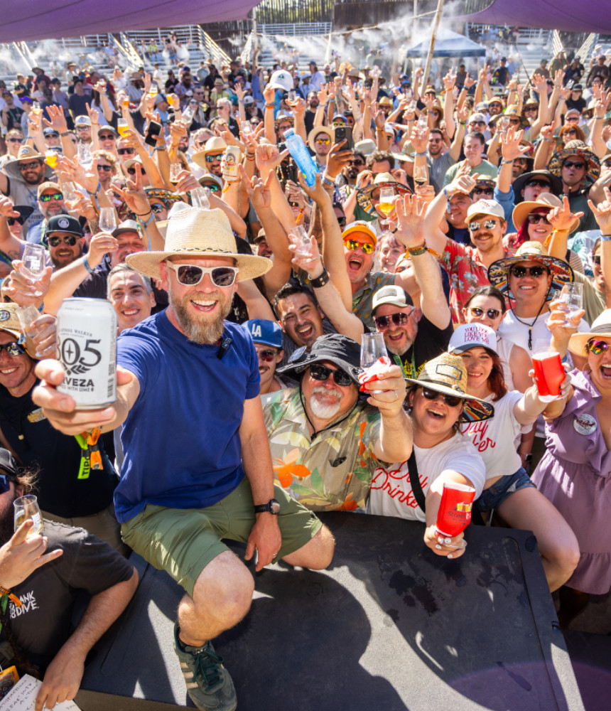 Group of people cheering with their arms in the air. Man front and center holding up a black and white 805 can on a stage. 