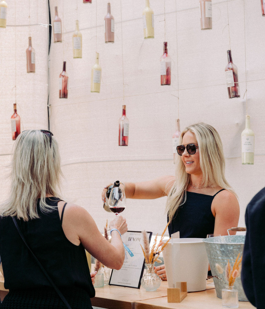 Two women in black shirts, one serving the other a glass of red wine with a variety of wine bottles hanging as decor in the background.